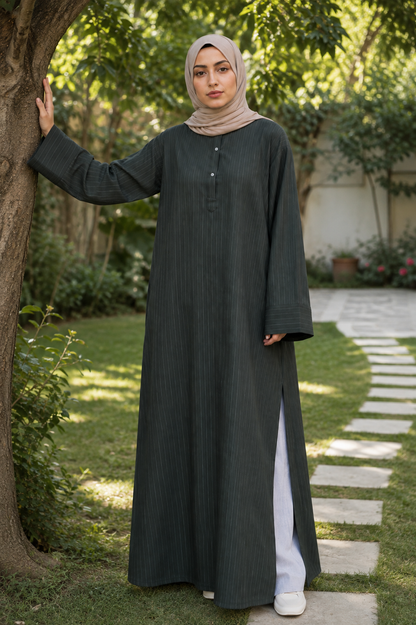 A woman wearing a dark charcoal gray long-sleeved abaya with a subtle vertical texture and a beige hijab, standing in a garden next to a tree.
