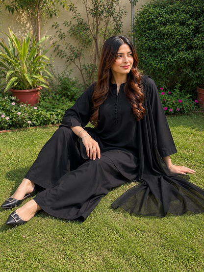A woman wearing a black traditional outfit with wide-leg pants and a draped cape-style top sits on a green lawn in a garden setting.